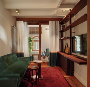 Una sala de estar con un sofá verde, una mesita de madera, una alfombra roja, un televisor sobre un mueble de madera y una ventana con cortinas.