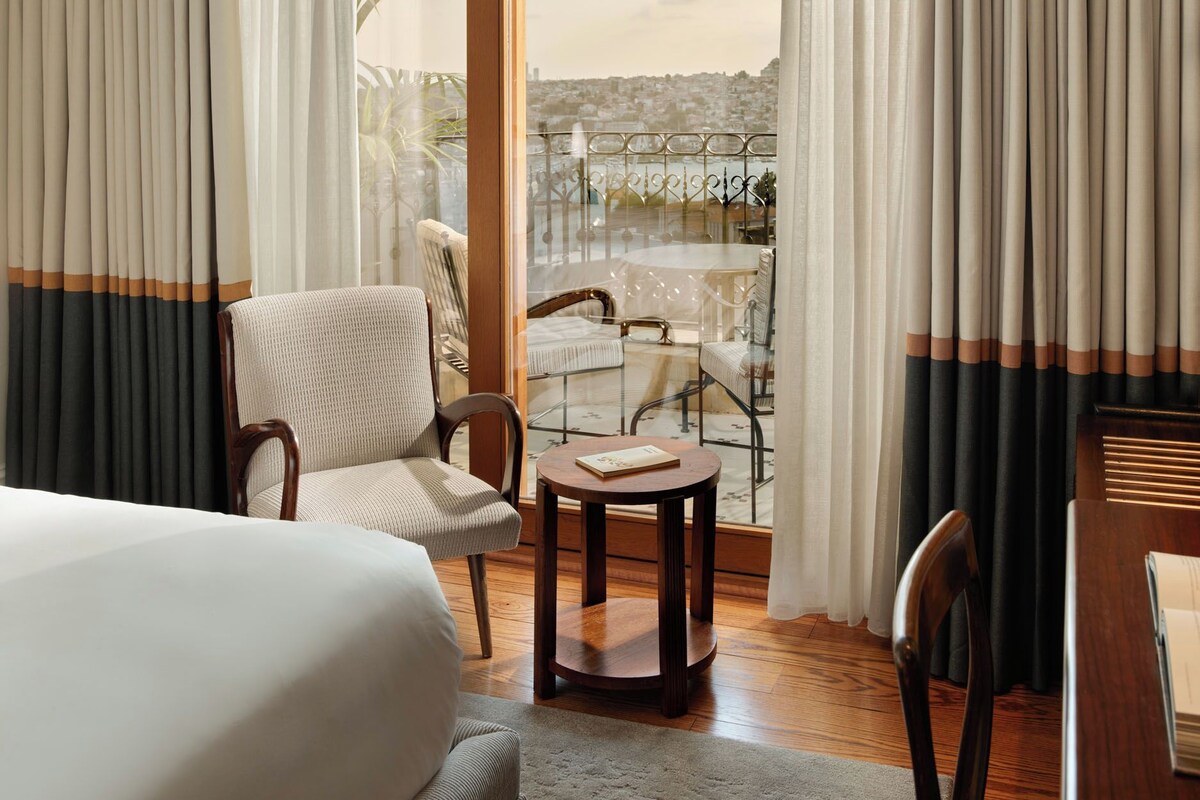 A hotel room with a bed, a round table, two chairs, a balcony with a view of a cityscape, and curtains.