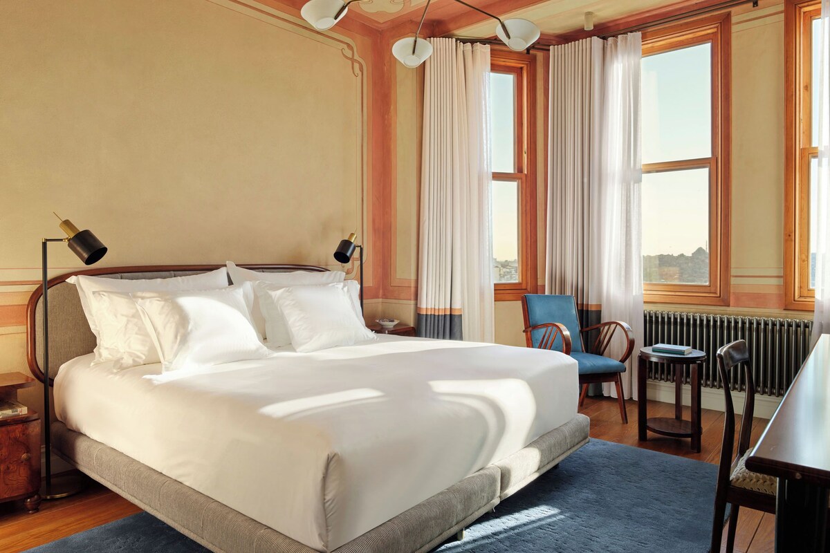 A hotel room with a large bed, two bedside lamps, a chair, a table, and a view of the city through the window.
