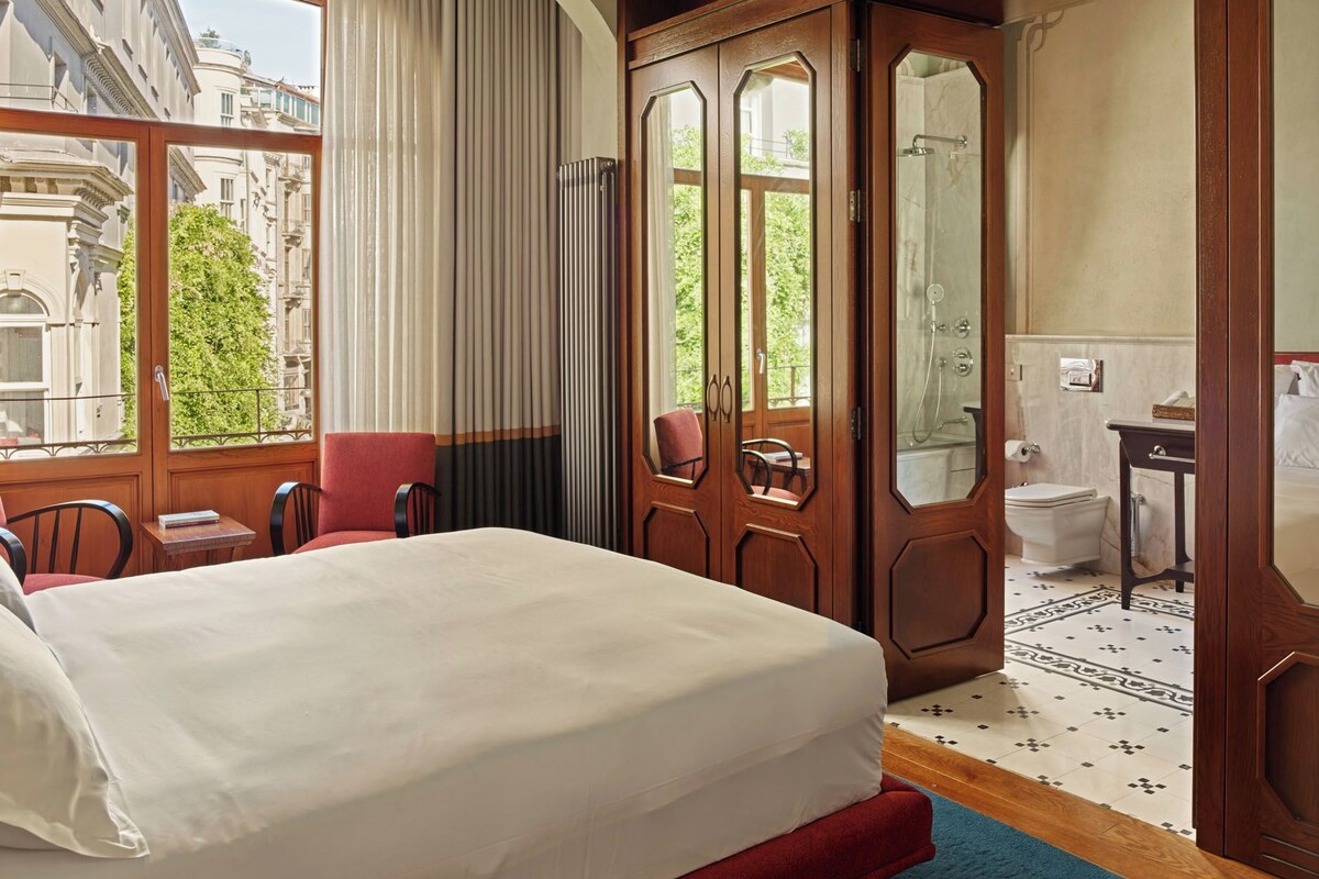 A hotel room with a large bed, a sitting area with a chair and a table, a bathroom with a shower and toilet, and a view of the city through a window.