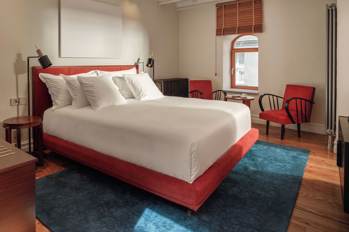 A hotel room with a large bed, a desk, a chair, and a window with blinds.