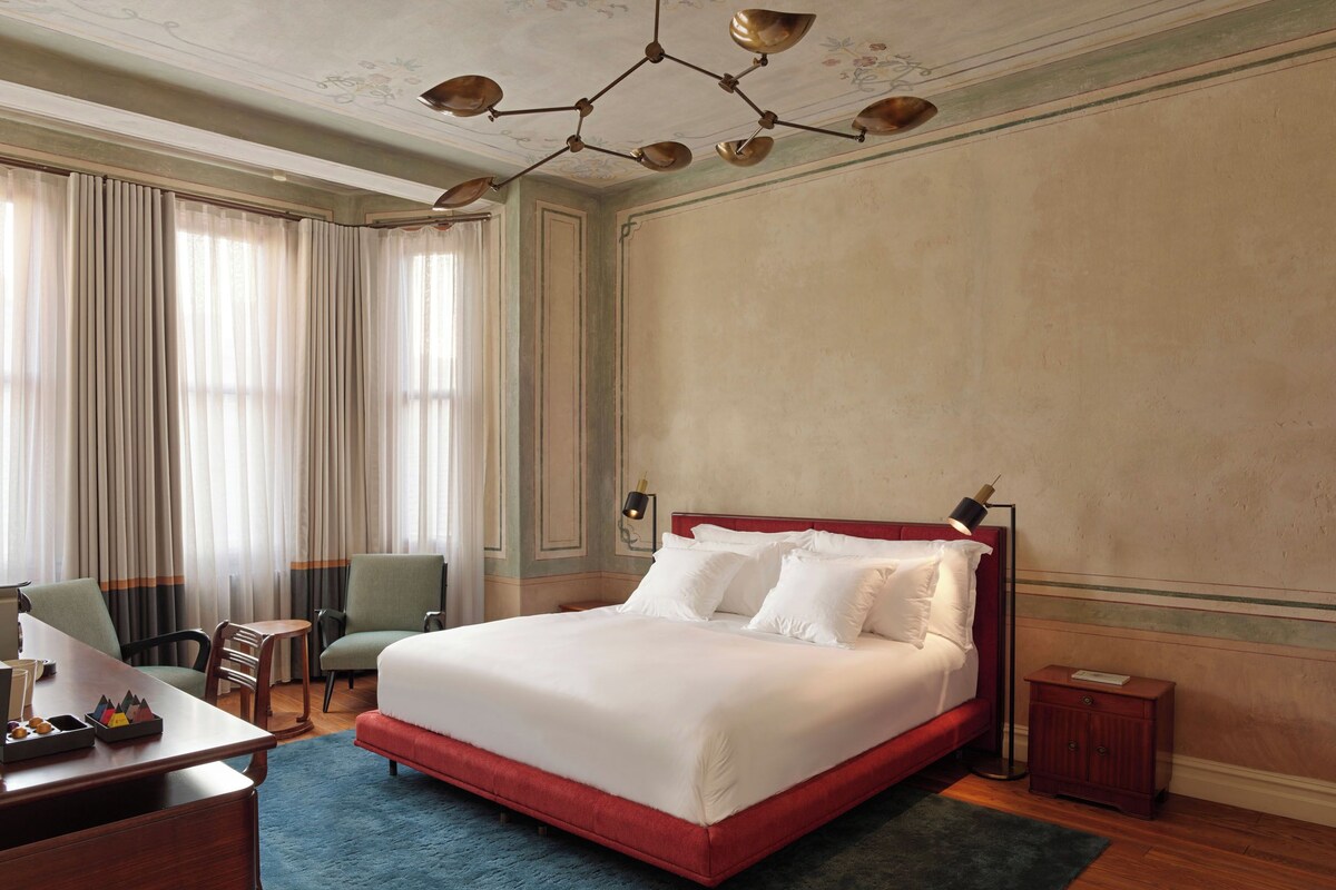 A hotel room with a large bed, a desk with a chair, a small table, and a nightstand.
