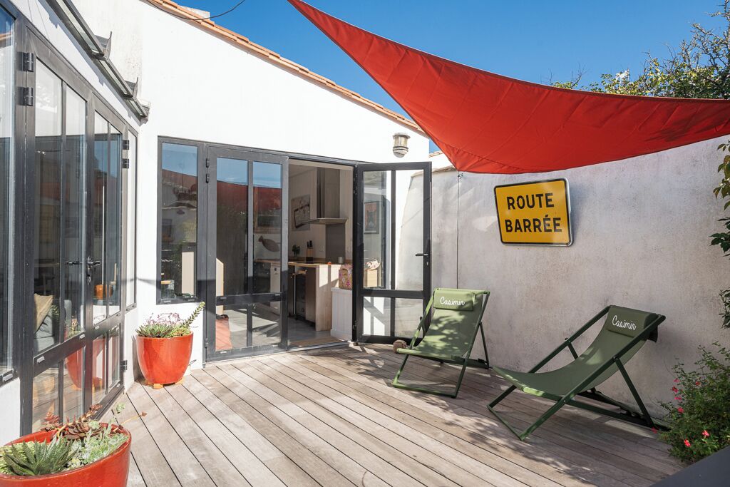 A covered outdoor area with a wooden deck, a red shade sail, and a sign reading 'ROUTE BARRÉE'.