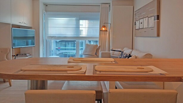 Un soggiorno moderno con un tavolo da pranzo in legno, sedie imbottite, un televisore a schermo piatto e una grande finestra con vista sulla città.