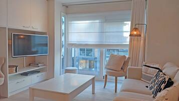 Een moderne woonkamer met een flatscreen televisie, een witte salontafel, een bank met decoratieve kussens en een fauteuil.