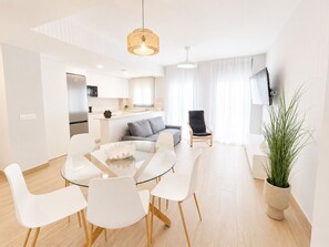 A modern living room with a glass dining table, white chairs, a grey sofa, a black armchair, and a potted plant. - Apartamento la Bodega con piscina (Conil de la Frontera)