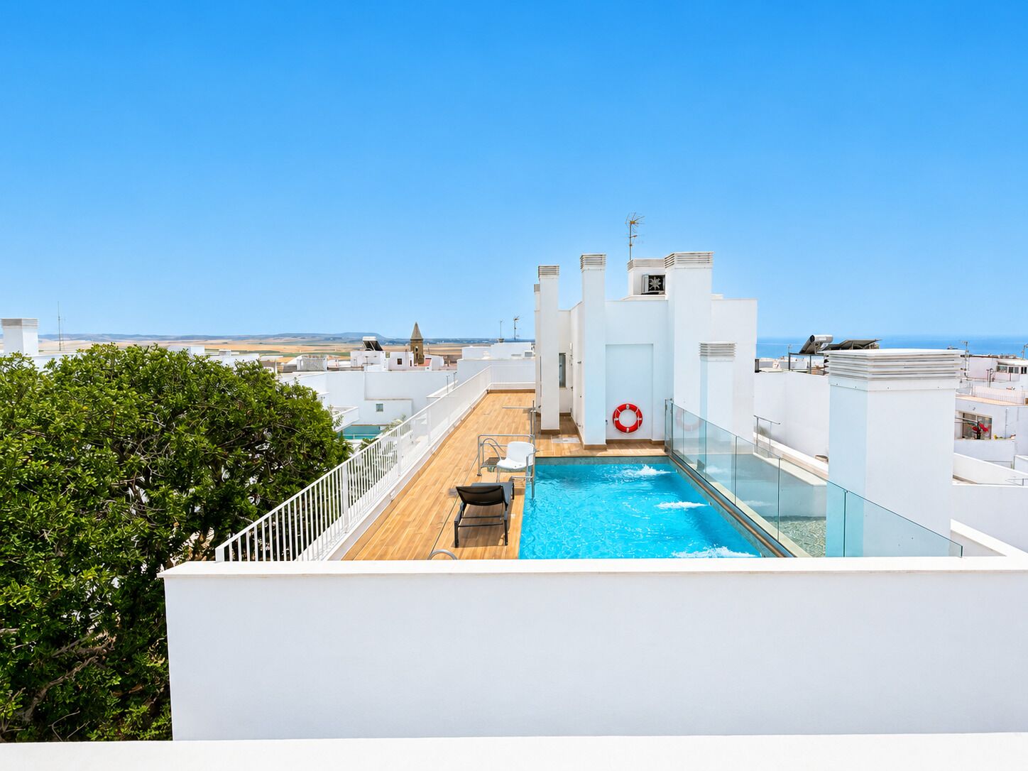 A rooftop pool with a clear view of the sea, surrounded by white buildings and a tree.