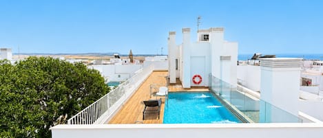 A rooftop pool with a clear view of the sea, surrounded by white buildings and a tree.