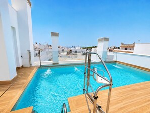 A rooftop swimming pool with a metal ladder and wooden deck, surrounded by white buildings. - Apartamento la Bodega con piscina (Conil de la Frontera)