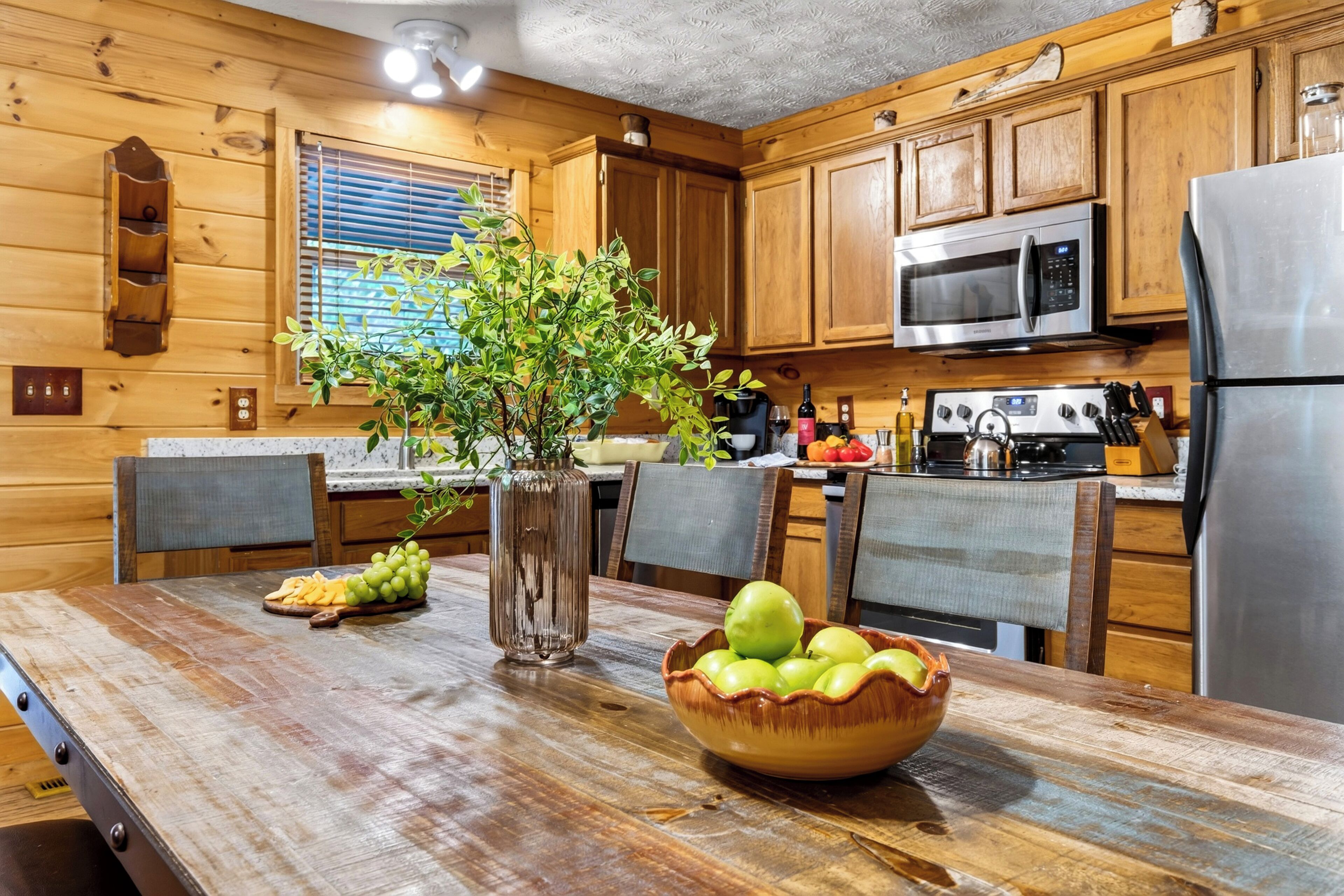 A wooden cabin interior with a kitchen area featuring wooden cabinets, a stainless steel refrigerator, a microwave, and a dining table with a vase of green plants and a bowl of apples.