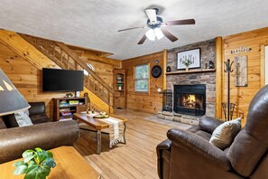 A cozy log cabin interior with a fireplace, leather furniture, a flat-screen TV, and a wooden staircase. - Hot Tub + Wraparound Decks + Arcade | Layered Ridge Views | Dream Chaser (Sevierville)