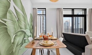 A modern dining area with a table set for two, a sofa, and large windows with a city view. - Blossom & Palm 1BR | designer Creek Harbour home (Dubai)