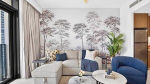 Una sala moderna con un sofá modular, una mesa de centro y un sillón azul. Se ve una ventana grande a la izquierda, una pared con un mural de un árbol, y un área de cocina a la derecha.