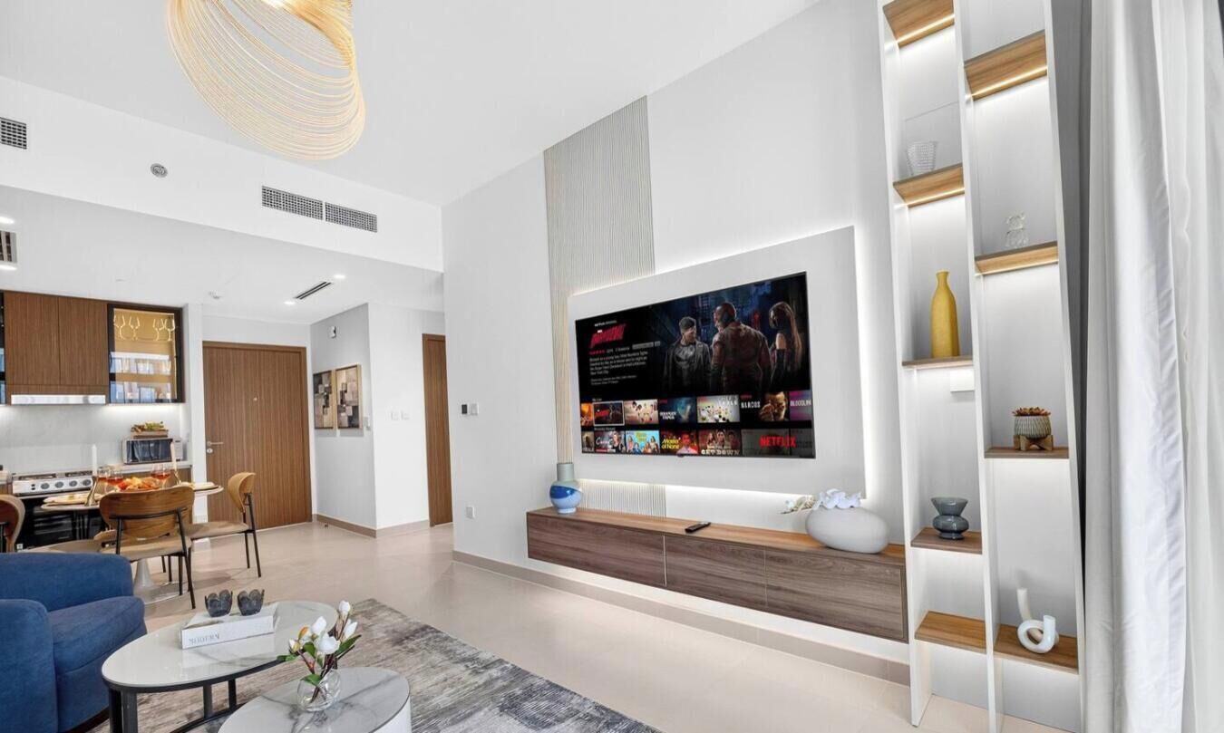 Ein modernes Wohnzimmer mit einem großen Flachbildfernseher an der Wand, einem hölzernen Konsolentisch und einer Sitzecke mit blauem Sofa und Couchtisch.