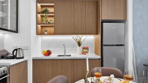 A modern kitchen with wooden cabinetry, a built-in shelf with plants, a refrigerator, and a dining area with a table set for two. - Feather & Stone 1BR | Creek Harbour skyline home (Dubai)