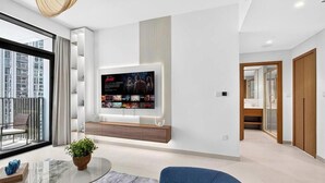 A modern living room with a large flat-screen TV mounted on the wall, a glass coffee table, and a view of the city through large windows. - Feather & Stone 1BR | Creek Harbour skyline home (Dubai)
