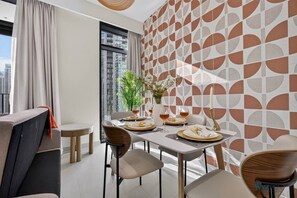 A modern dining area with a table set for four, a potted plant, and a city view through large windows. - Copper & Clay 1BR | warm Creek Harbour home (Dubai)
