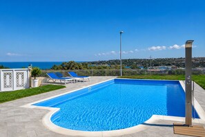 Ein rechteckiger Swimmingpool mit geschwungener Kante, umgeben von einem weißen Zaun und zwei blauen Liegestühlen.