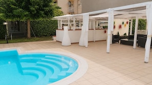 A swimming pool with a tiled deck, a covered seating area with tables and chairs, and a building in the background. - Vacation Home in Arenella with 4 bedrooms sleeps 11 (Arenella)