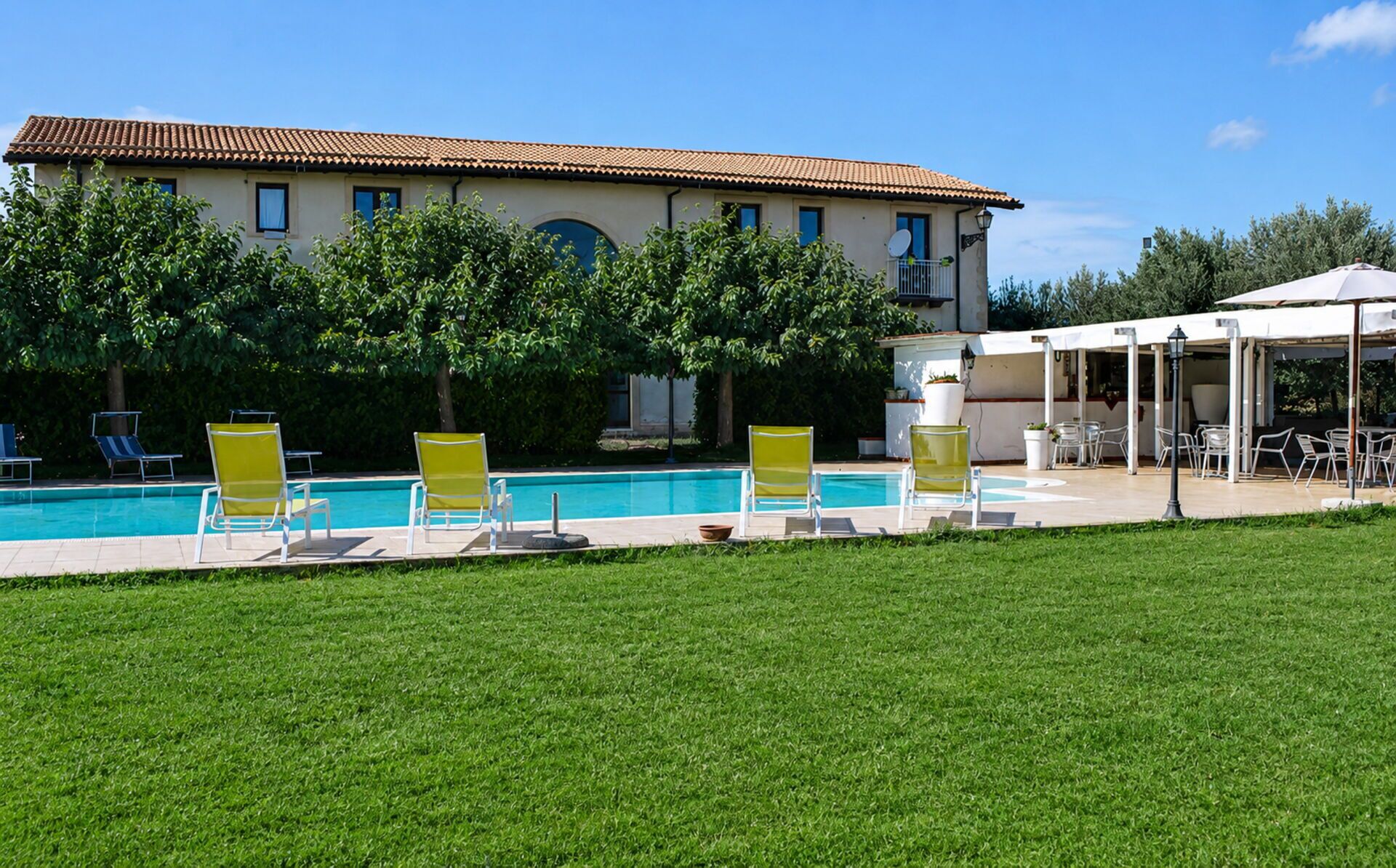 A pool area with lounge chairs, a building with a tiled roof, and a covered outdoor seating area.