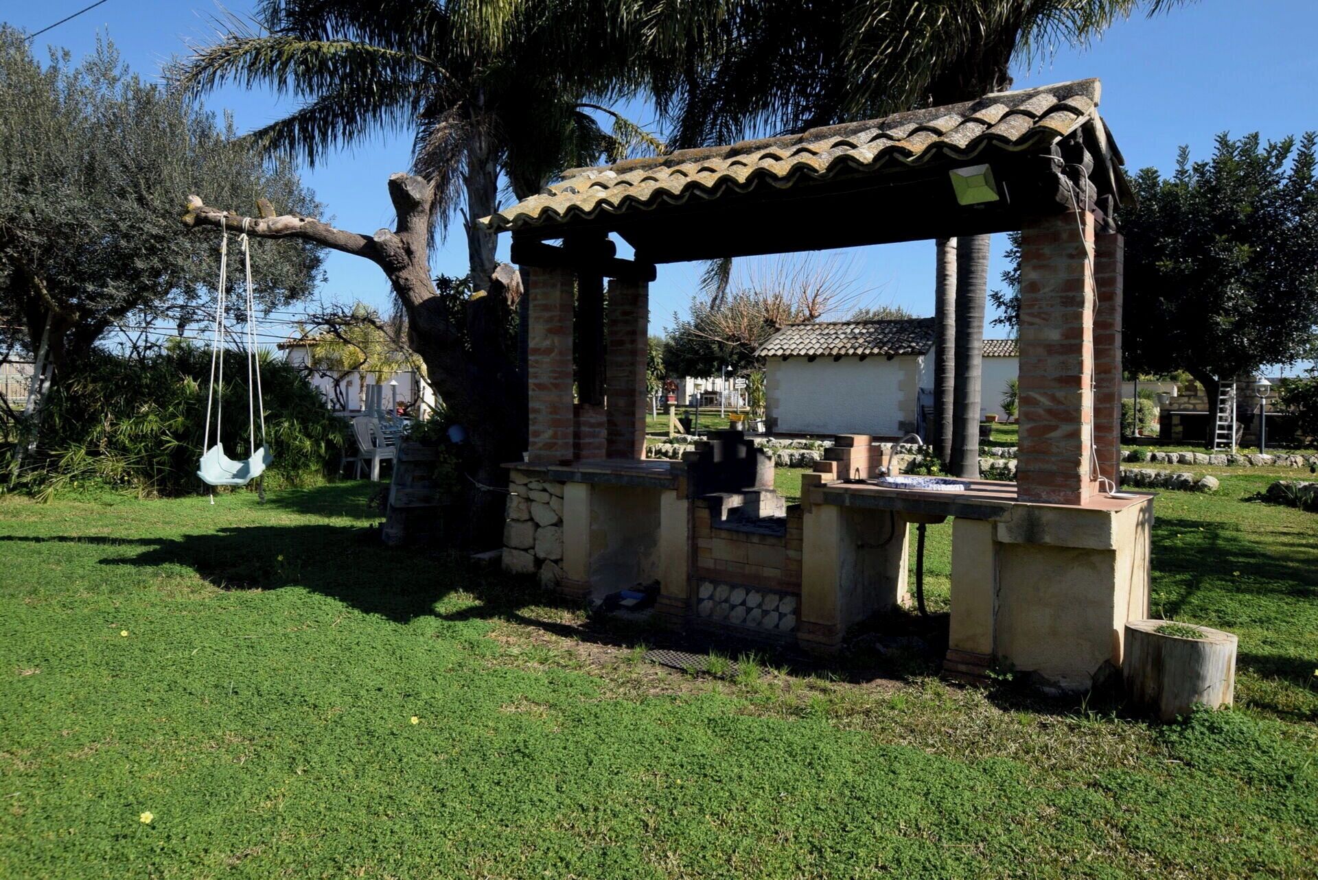 A rustic outdoor seating area with a swing, stone pillars, and a tiled roof.