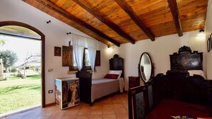 A room with a wooden ceiling, a bed with a headboard, a bedside table, a mirror, and a view of a garden.