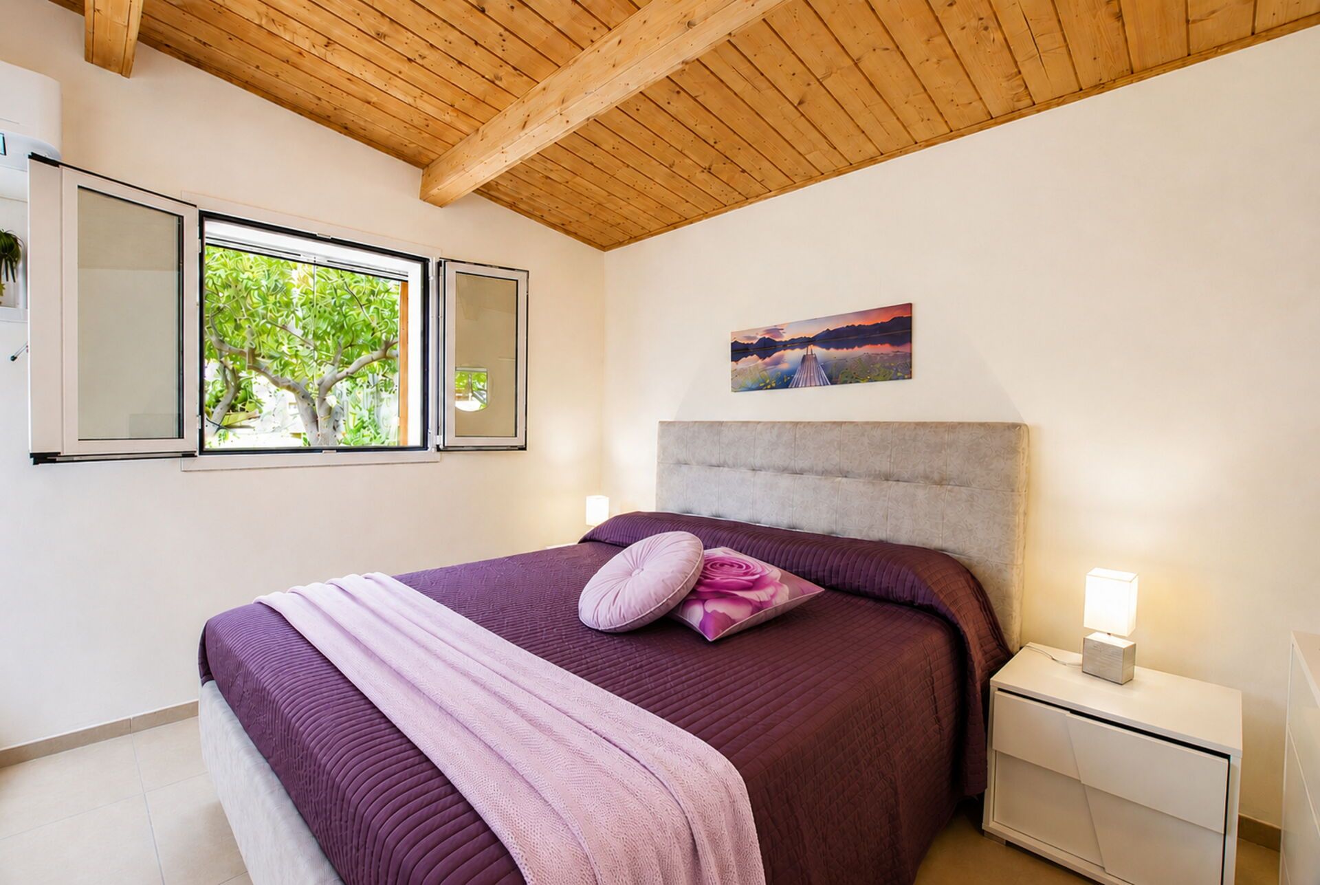 A bedroom with a bed, nightstands, a painting, and a window with a view of trees.