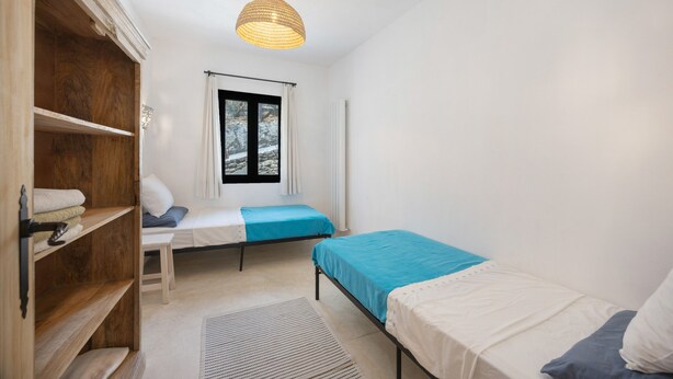 A small, well-lit bedroom with a single bed, a wooden shelf with towels, a window with a view of trees, and a hanging light fixture.