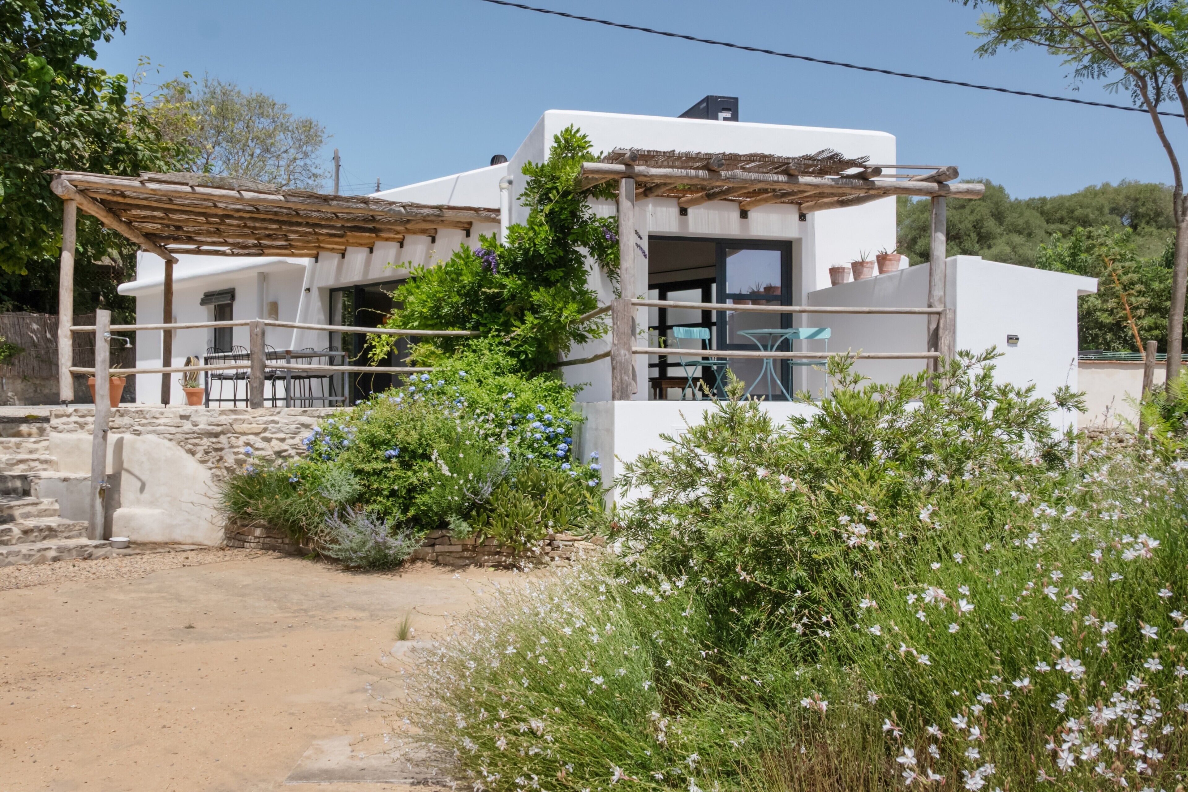 Un edificio blanco con una pérgola de madera, asientos al aire libre y un muro de piedra.