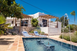 Zona de piscina con sillas de descanso, una pérgola y una escalera de piedra que conduce a un edificio blanco.