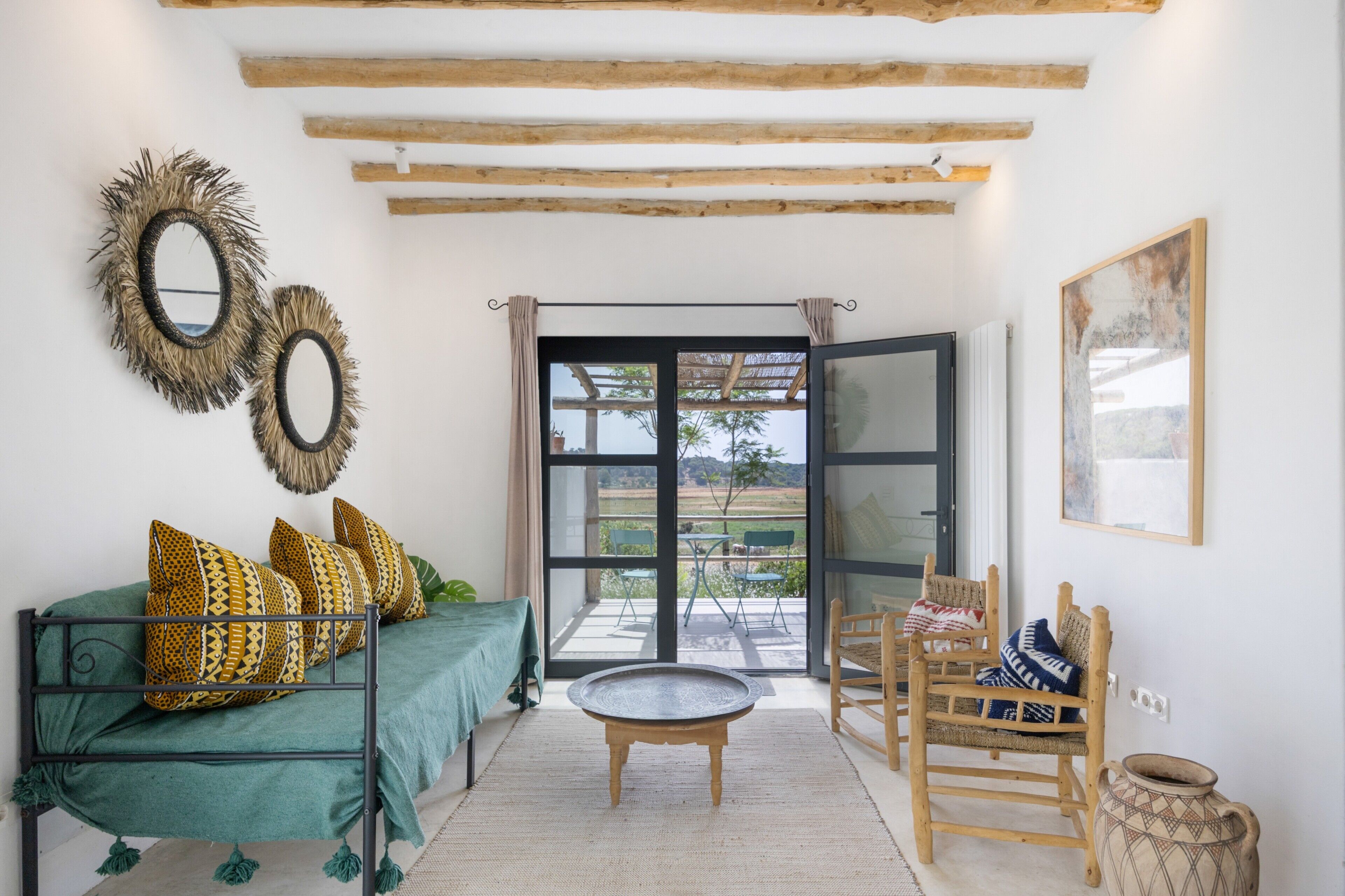 A room with a green sofa, patterned cushions, a round coffee table, a wooden chair, a woven basket, and a large window with a view of the outdoors.