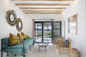A room with a green sofa, patterned cushions, a round coffee table, a wooden chair, a woven basket, and a large window with a view of the outdoors.