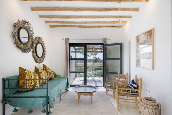 Una habitación con sofá verde, cojines estampados, una mesa de centro redonda, una silla de madera, una cesta tejida y un ventanal con vistas al exterior.