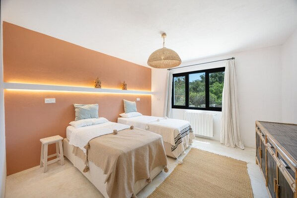 A bedroom with two beds, a small table, a hanging light fixture, and a window with a view of greenery.