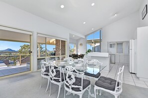 A modern dining area with a glass table and white chairs, a kitchen with white appliances, and a view of the outdoors. - Bella on Canomii Family Beach House in Nelson Bay (Nelson Bay)