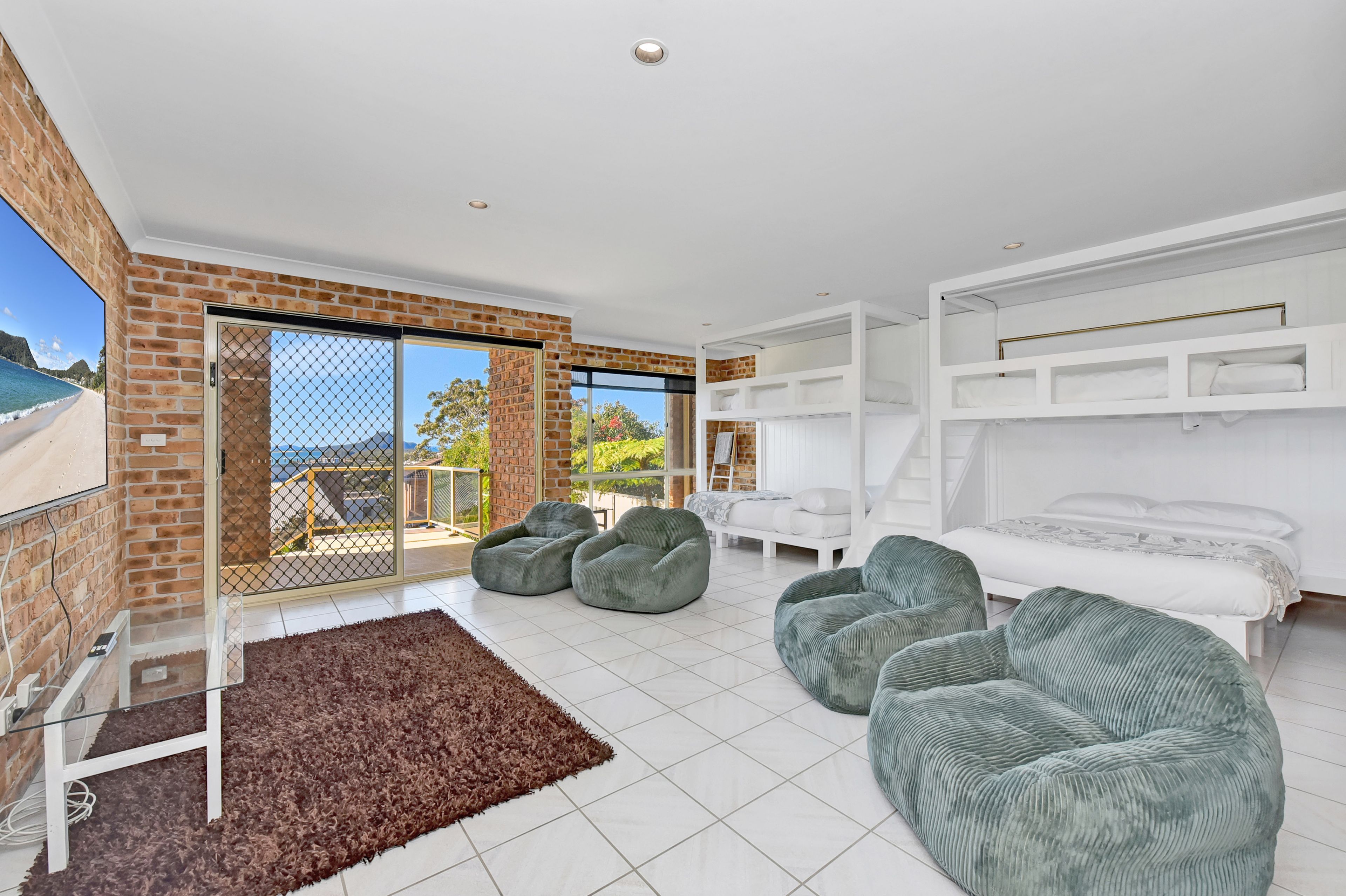 A modern living room with a large sliding glass door leading to a balcony, a white bed with a bunk bed above, and two green bean bag chairs.