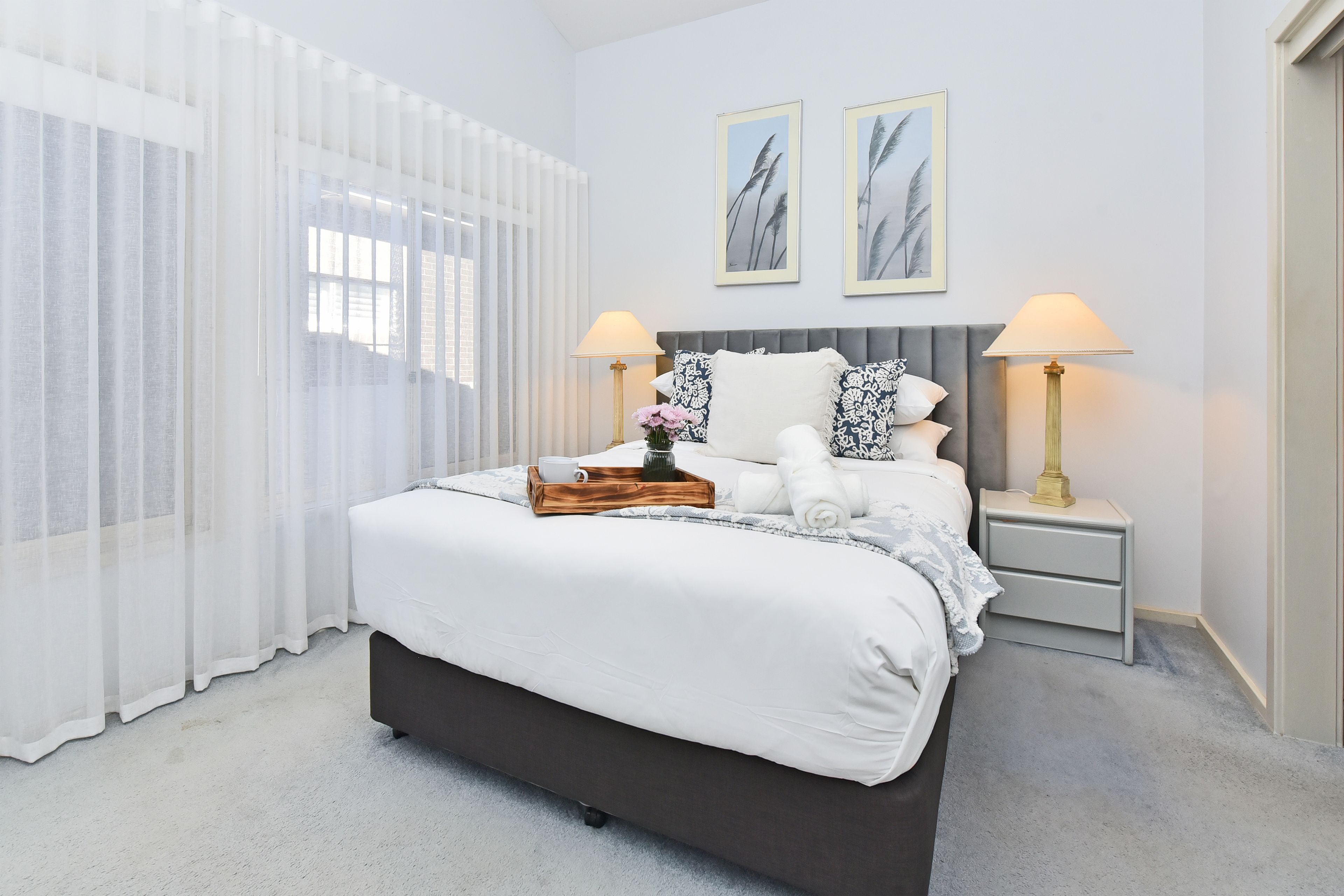 A bedroom with a bed, two bedside tables, a window with sheer curtains, and two wall-mounted picture frames.