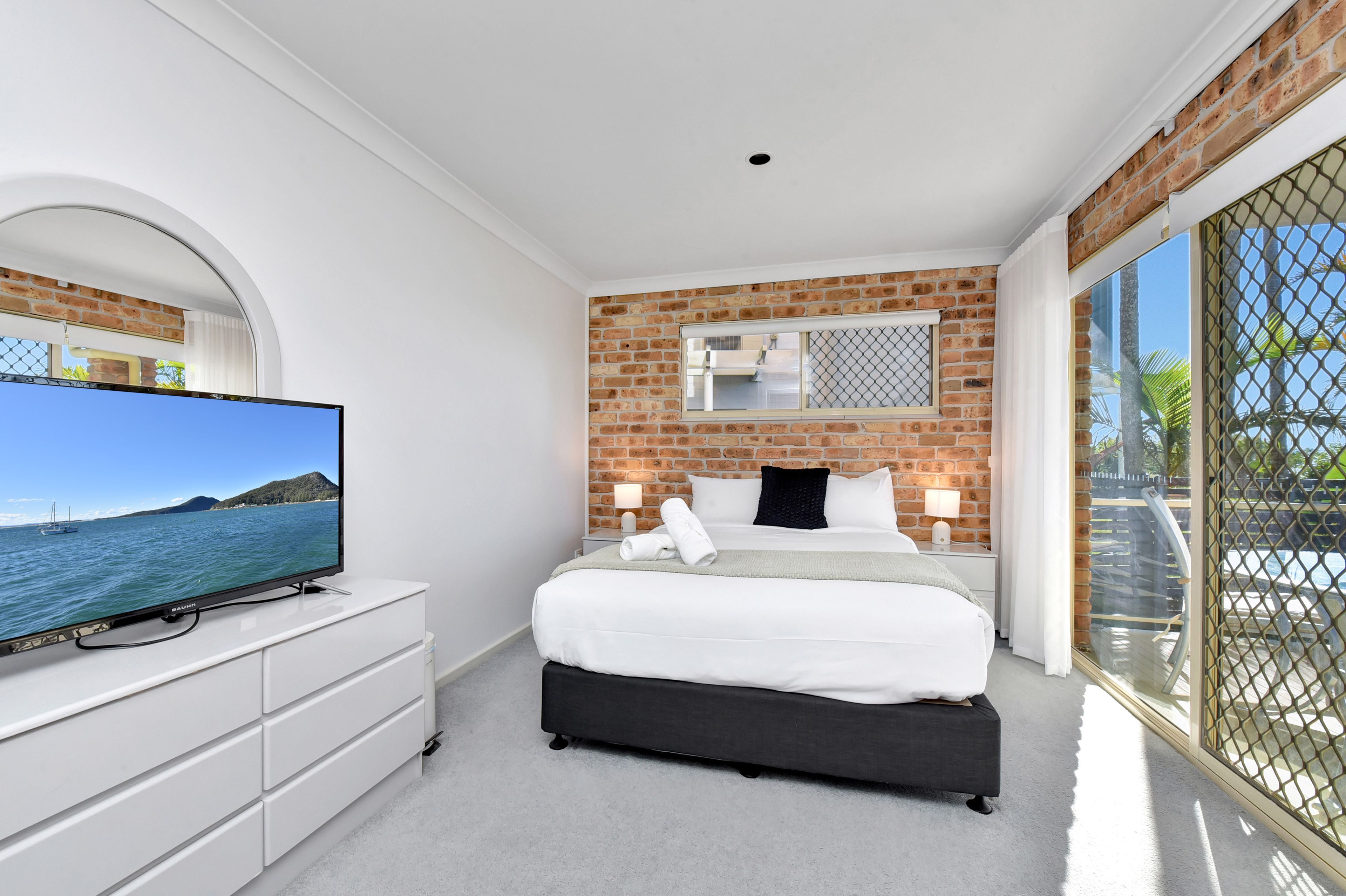 A hotel room with a large TV, a bed with white and black bedding, a brick accent wall, and a balcony with a view of the sea.