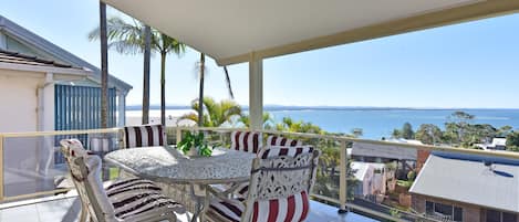 Un balcon avec une table et des chaises, donnant sur une étendue d’eau et sur une terre lointaine.