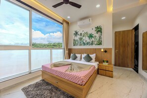 Ein Schlafzimmer mit großem Fenster, Deckenventilator, einem Bett mit gemusterten Textilien, einem Nachttisch und einer Wandklimaanlage.