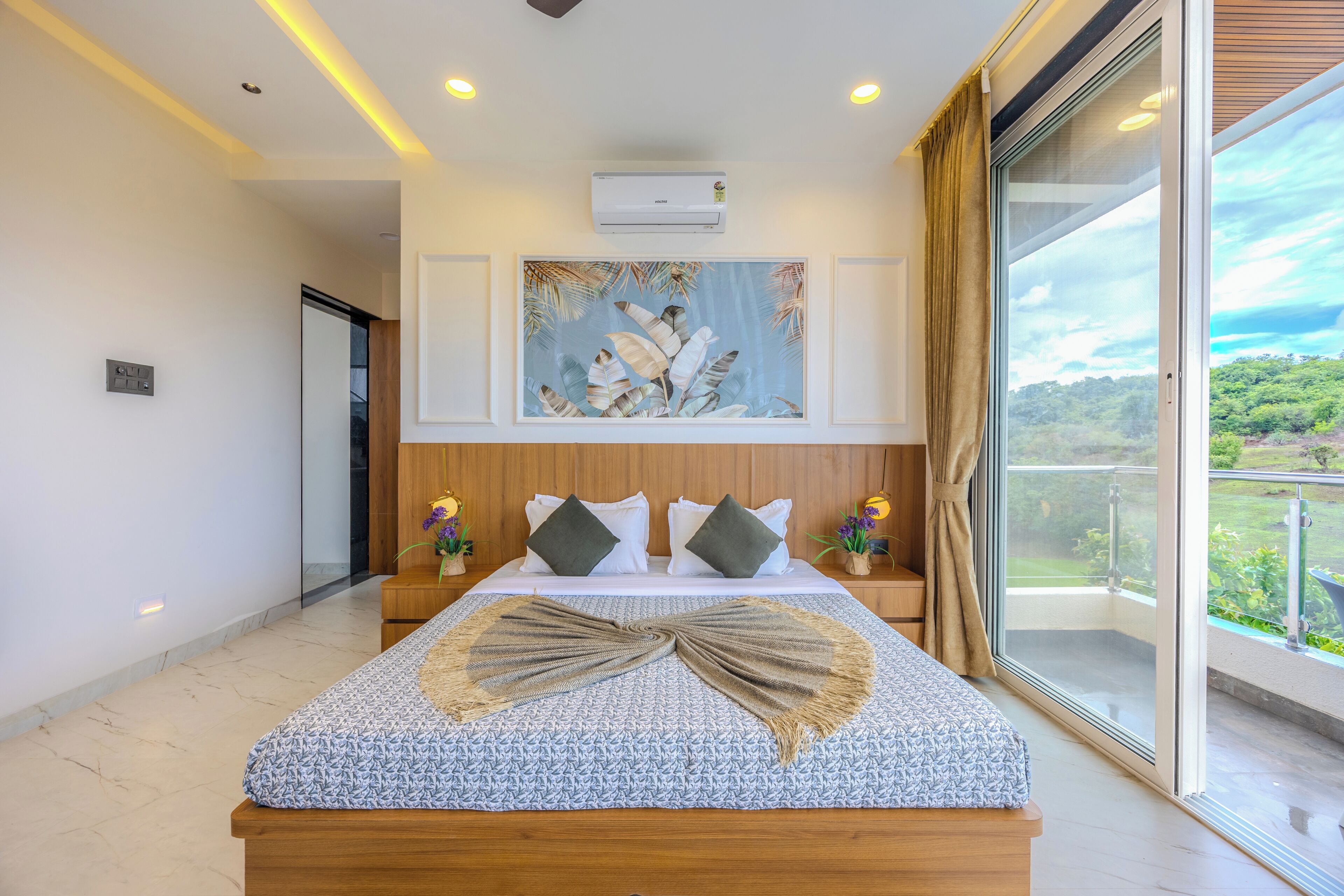 Une chambre moderne avec un grand lit, un climatiseur fixé au plafond, une œuvre d’art tropical encadrée et une vue sur la verdure par une grande fenêtre.