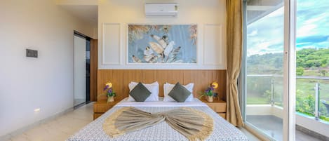 A modern bedroom with a large bed, a ceiling-mounted air conditioner, a framed tropical artwork, and a view of greenery through a large window.