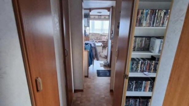 A narrow hallway with a checkered floor leading to a room with a bed and a window.