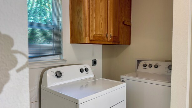 A laundry room with a front-loading washer and dryer, wooden cabinets above, and a window with blinds.