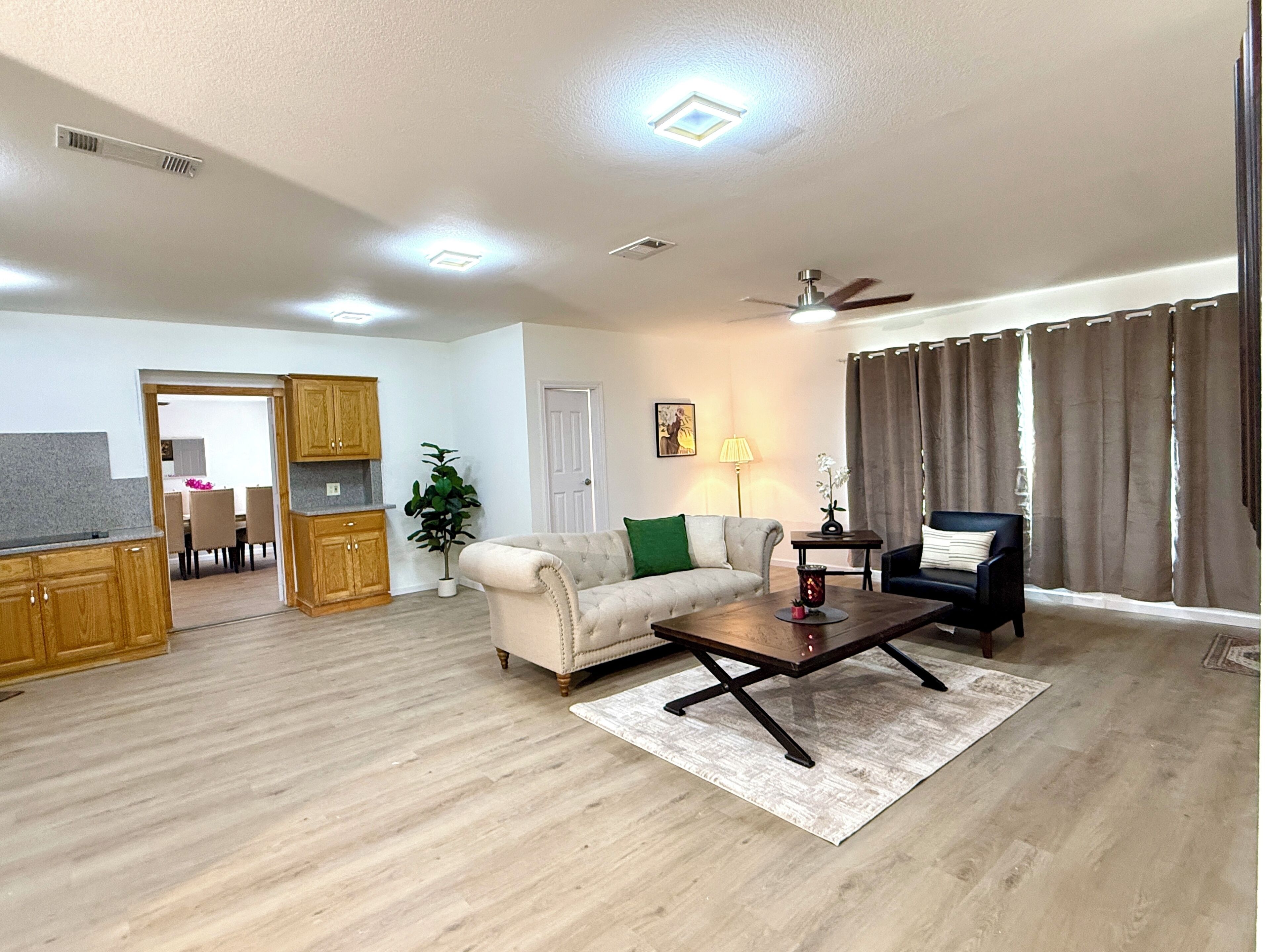 A living room with a sofa, a coffee table, a chair, a dining area with a table and chairs, and a kitchen with wooden cabinets.