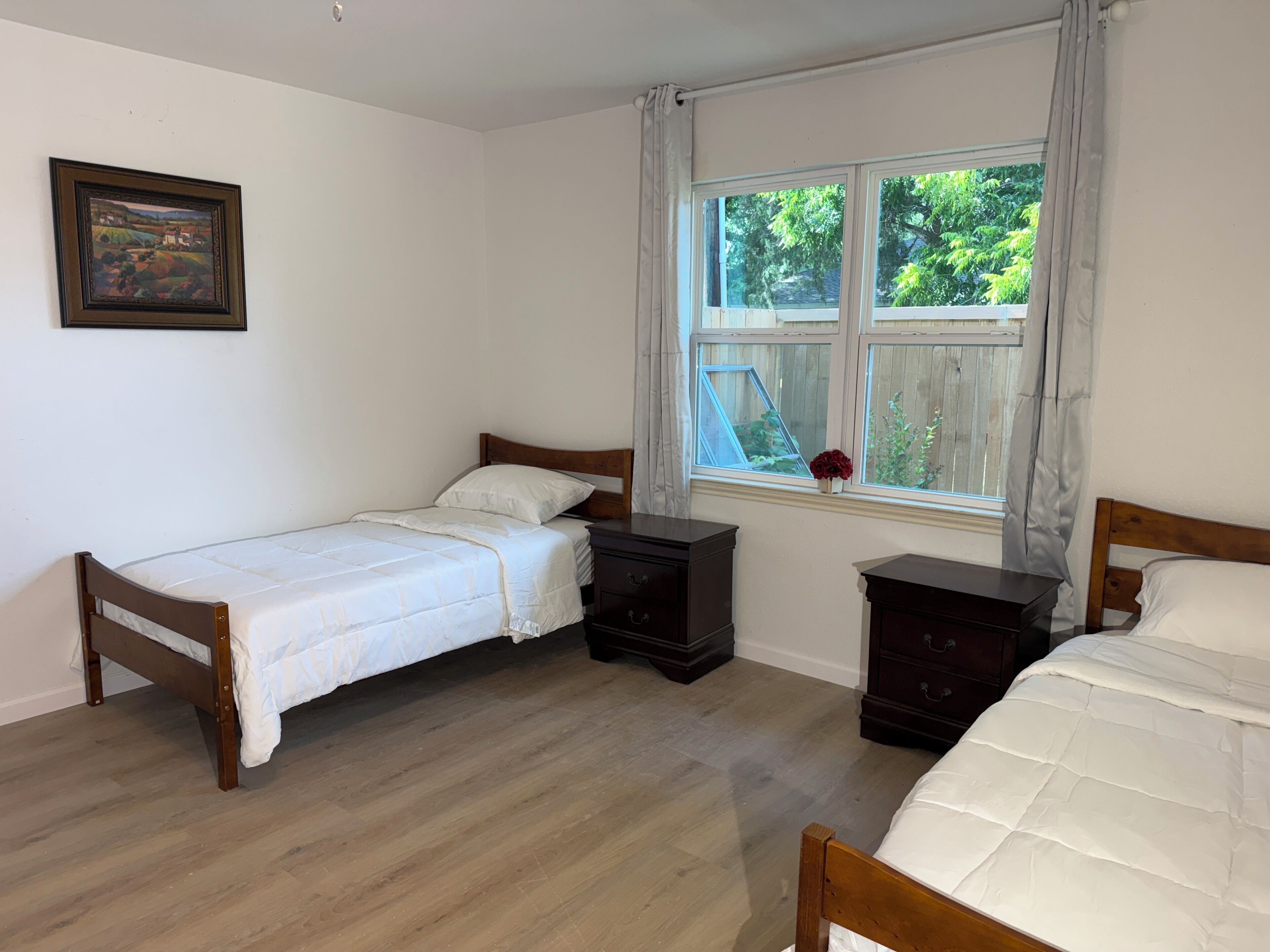 Een slaapkamer met twee eenpersoonsbedden, een houten dressoir, een nachtkastje, een raam met uitzicht op groen en een lijst met een afbeelding aan de muur.
