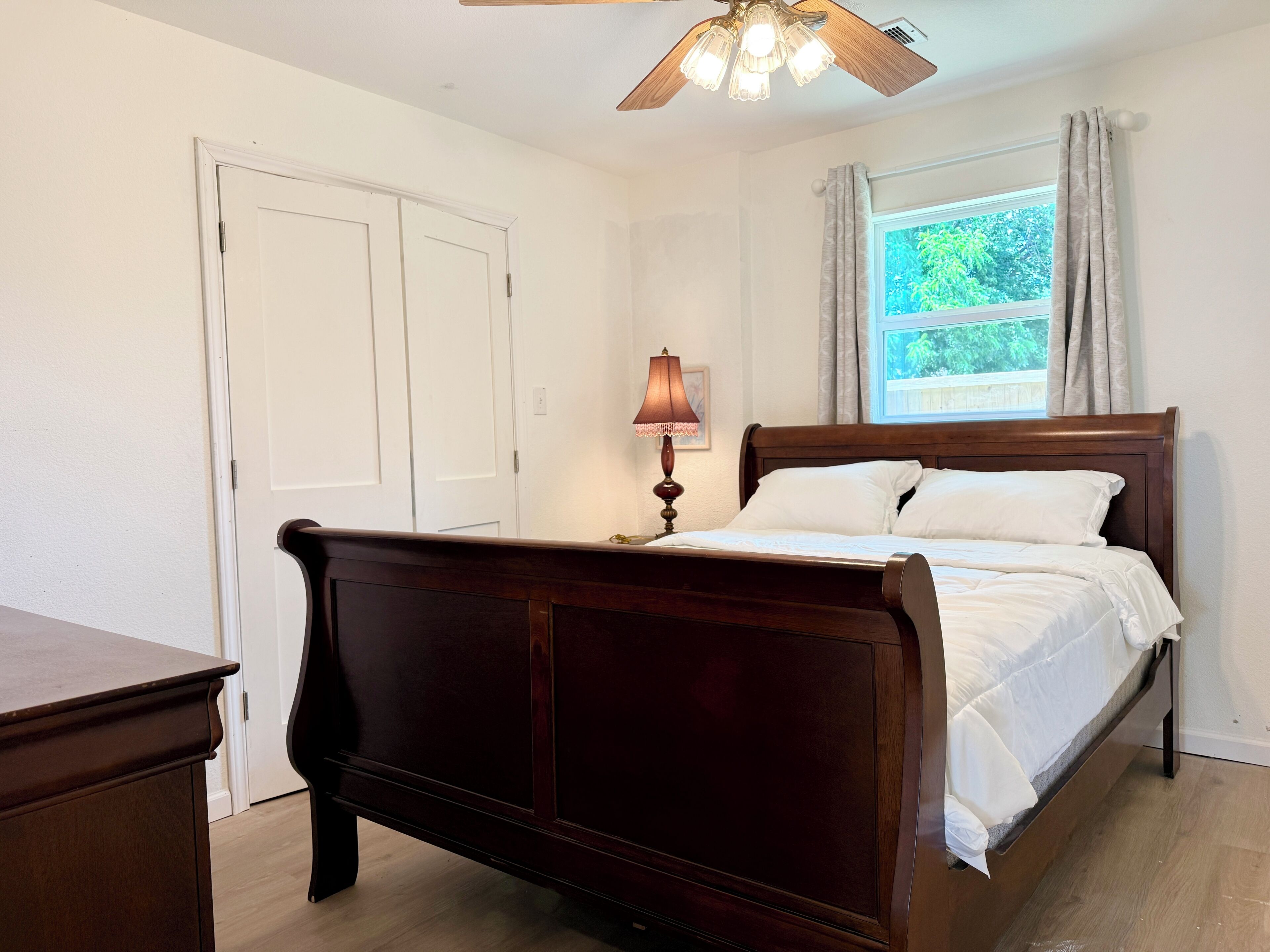 A bedroom with a wooden bed, a nightstand, a lamp, a ceiling fan, and a window with curtains.