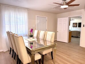 Un coin repas avec une table à plateau en verre, des chaises recouvertes d’un tissu beige et un ventilateur de plafond.
