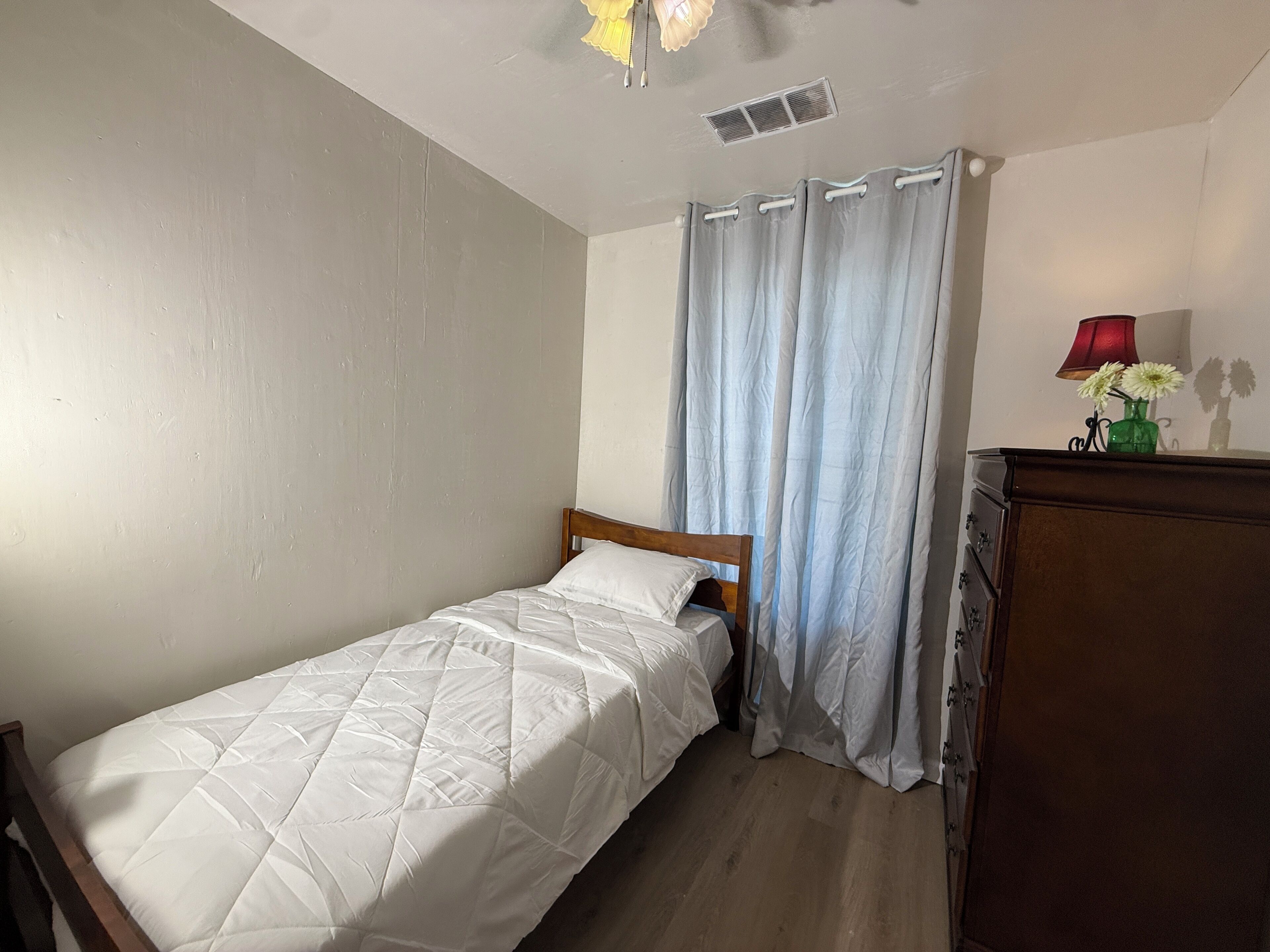 A single bed with a wooden headboard, a small dresser, a lamp, and a window with light blue curtains.