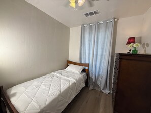 Un lit simple avec une tête de lit en bois, une petite commode, une lampe et une fenêtre avec des rideaux bleu clair.